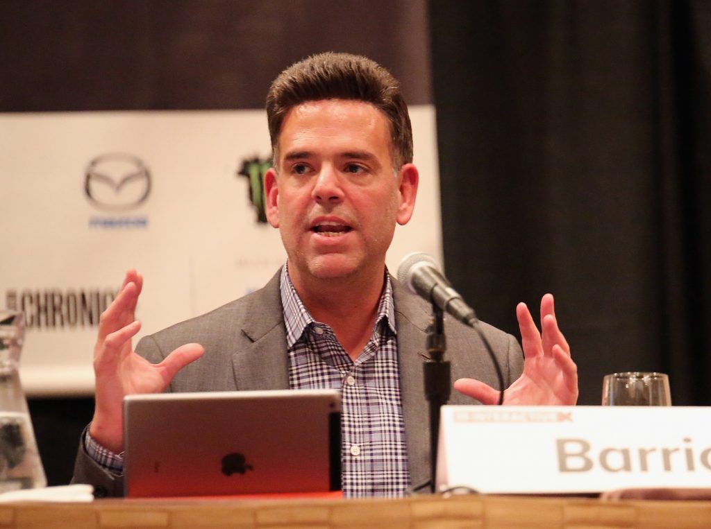 A man seated at a table gestures with his hands while speaking.