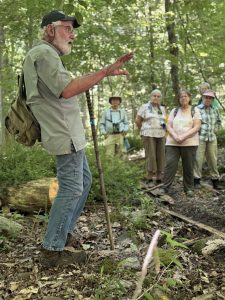 An older man with a hat, backpack, and a walking stick speaks to a group of five people in a forest