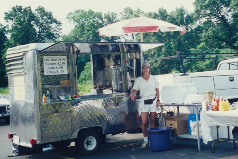 A vintage photo of a woman dressed in white with black phanny pack, standing next to a hot dog cart in a parking lot.