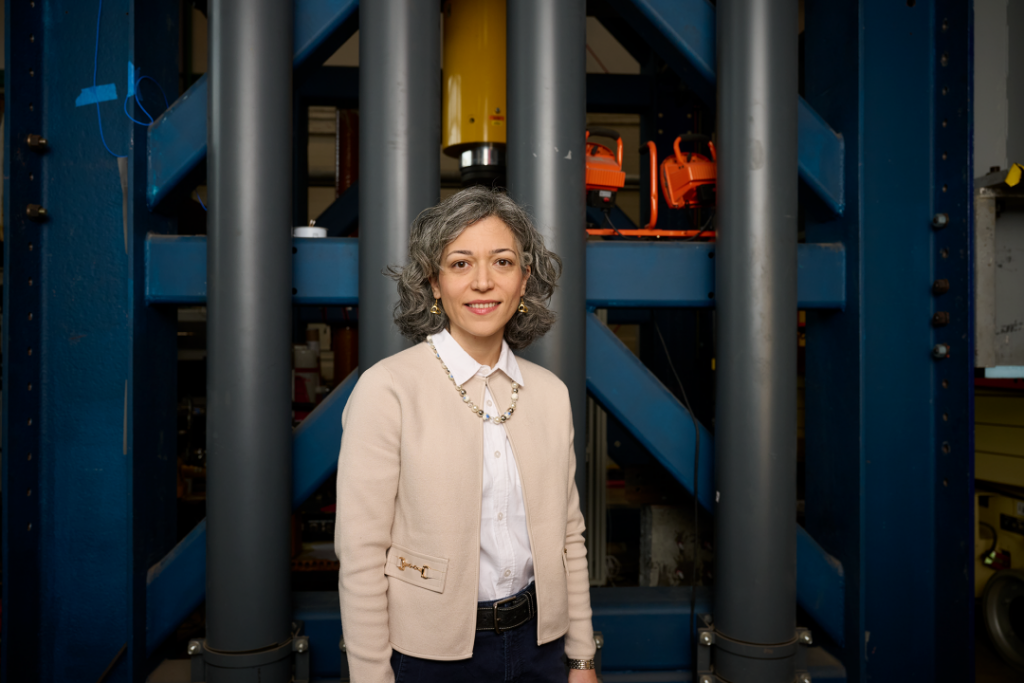 Motaref in khaki cardigan and black pants stands in front of a large metal structure.