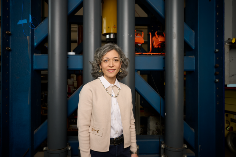 Motaref in khaki cardigan and black pants stands in front of a large metal structure.