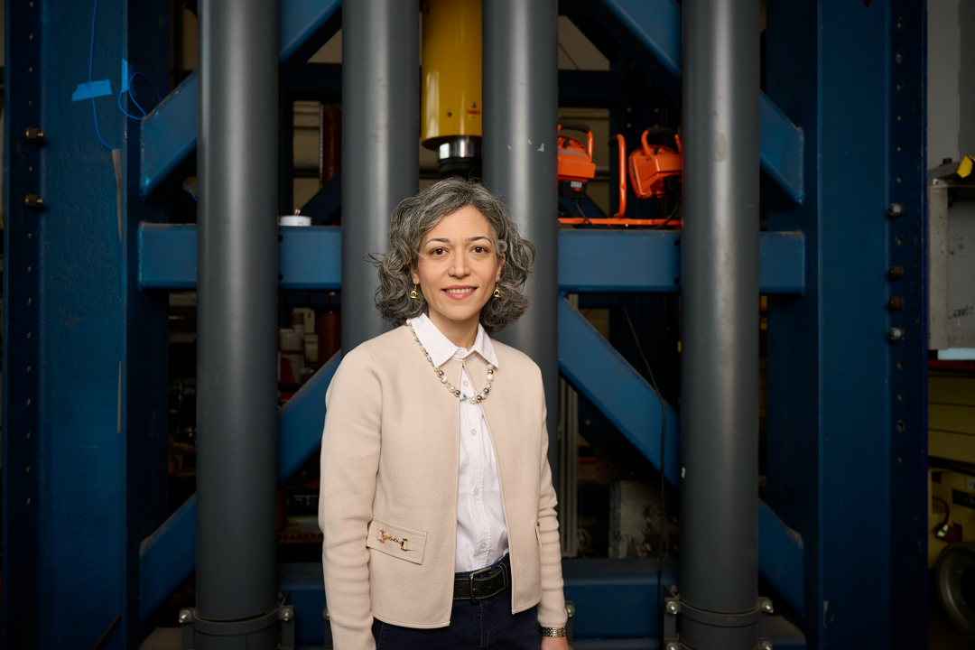 Motaref in khaki cardigan and black pants stands in front of a large metal structure.