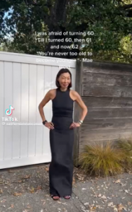 An older woman dressed in a sleeveless floor-length black dress stands with her hands on her hips.