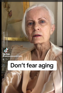 An older woman dressed in a satin shirt looks into the camera as the words