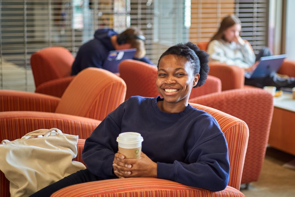 Moyo, woman in blue, sits in a soft orange armchair, holding a coffee cup. She is smiling and appears to be enjoying her time in the cozy atmosphere.