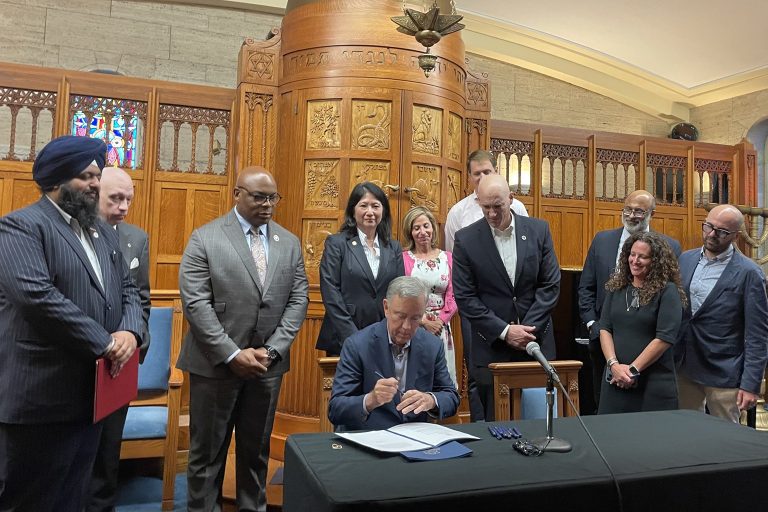 Ten people stand behind a man who is sitting at table signing a piece of paper.