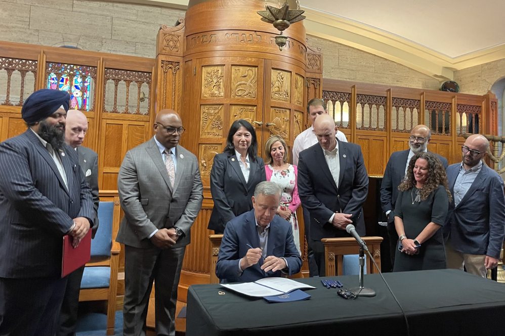 Ten people stand behind a man who is sitting at table signing a piece of paper.