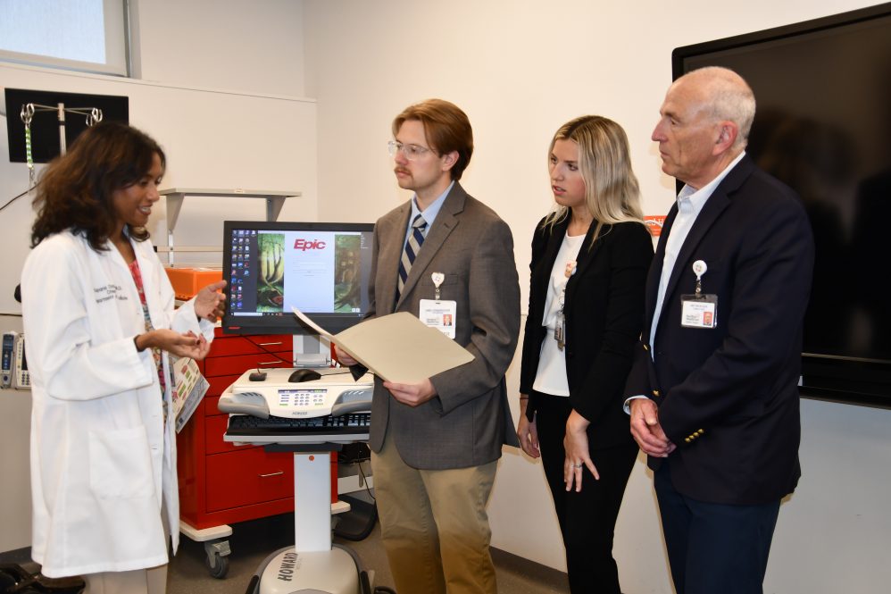 A doctor in a white lab coat talks to two students and a professor in front of a computer in a hospital room.