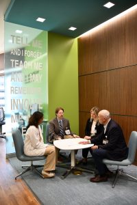 Four people sit at a table in a room with a high ceiling and glass wall.