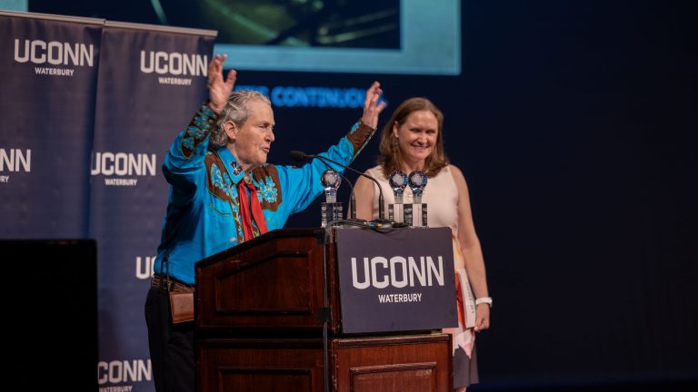 A woman in a Western-style shirt stands at a podium, addressing a crowd.