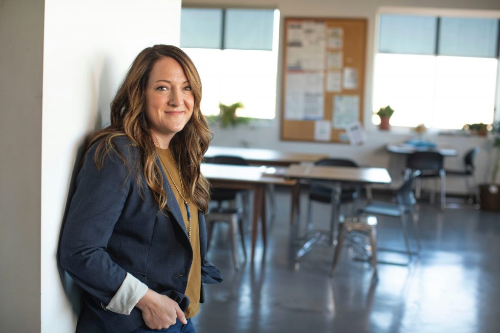 A woman in a blazer leaning against the wall of a classroom