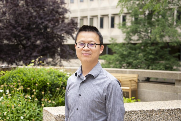 A man with glasses and a button-down shirt poses outside in front of bushes and trees with a building in the background.