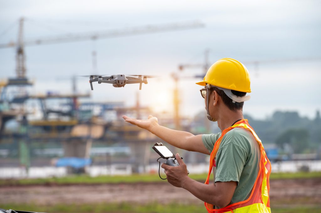 A worker in a safety helmet and neon visibility vest standing in an industrial zone. He holds a controller for a drone that has just taken off from his hand