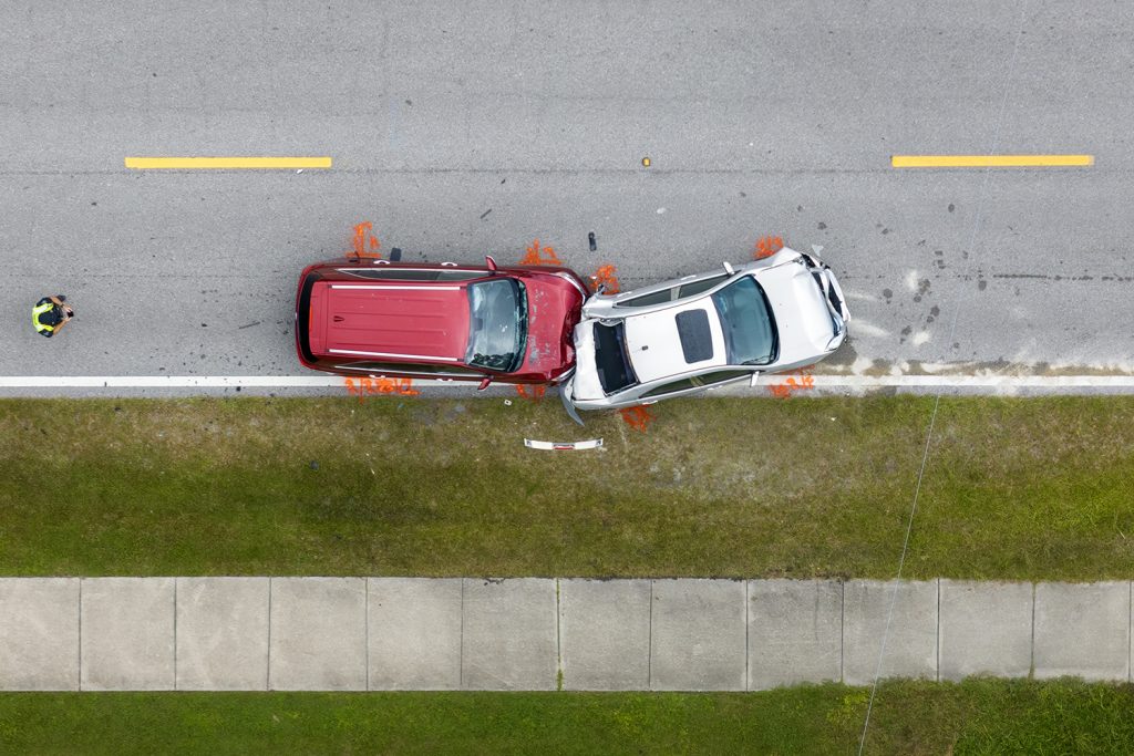 An aerial shot captures the aftermath of a collision between a red SUV and a silver car, the vehicles angled against each other on a grey asphalt road. Debris litters the roadside, marked by orange paint. A person in a bright vest stands off to the side.