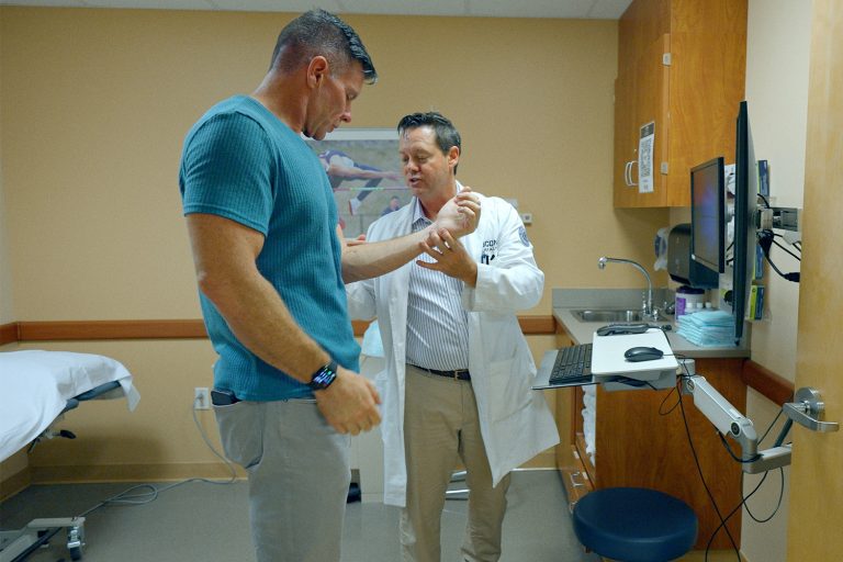 A patient in a blue shirt stands with a doctor examining his wrist