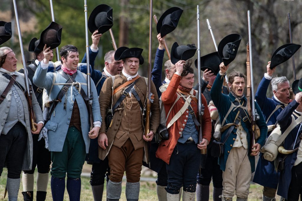 Revolutionary War reenactors stand in a line holding their hats above their heads.