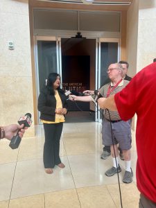 A woman dressed in black pants, black suit jacket, and yellow shirt talks to members of the media holding microphones in her direction.