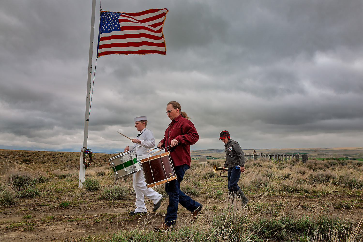two men walking with drums and a boy walking with a bugle by a flagpole