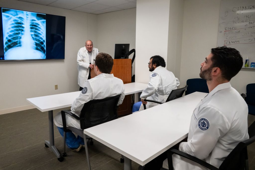 Man at podium, X-ray image to his right, speaking to three seated men
