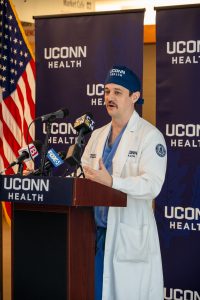 A doctor in a white coat and surgical hat speaking at a podium