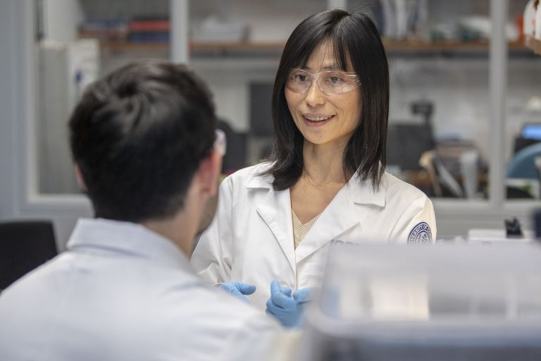 Woman with black hair in lab coat doing an experiment with a male student