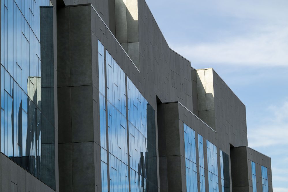 A portion of a grey building, with large windows reflecting the blue sky.