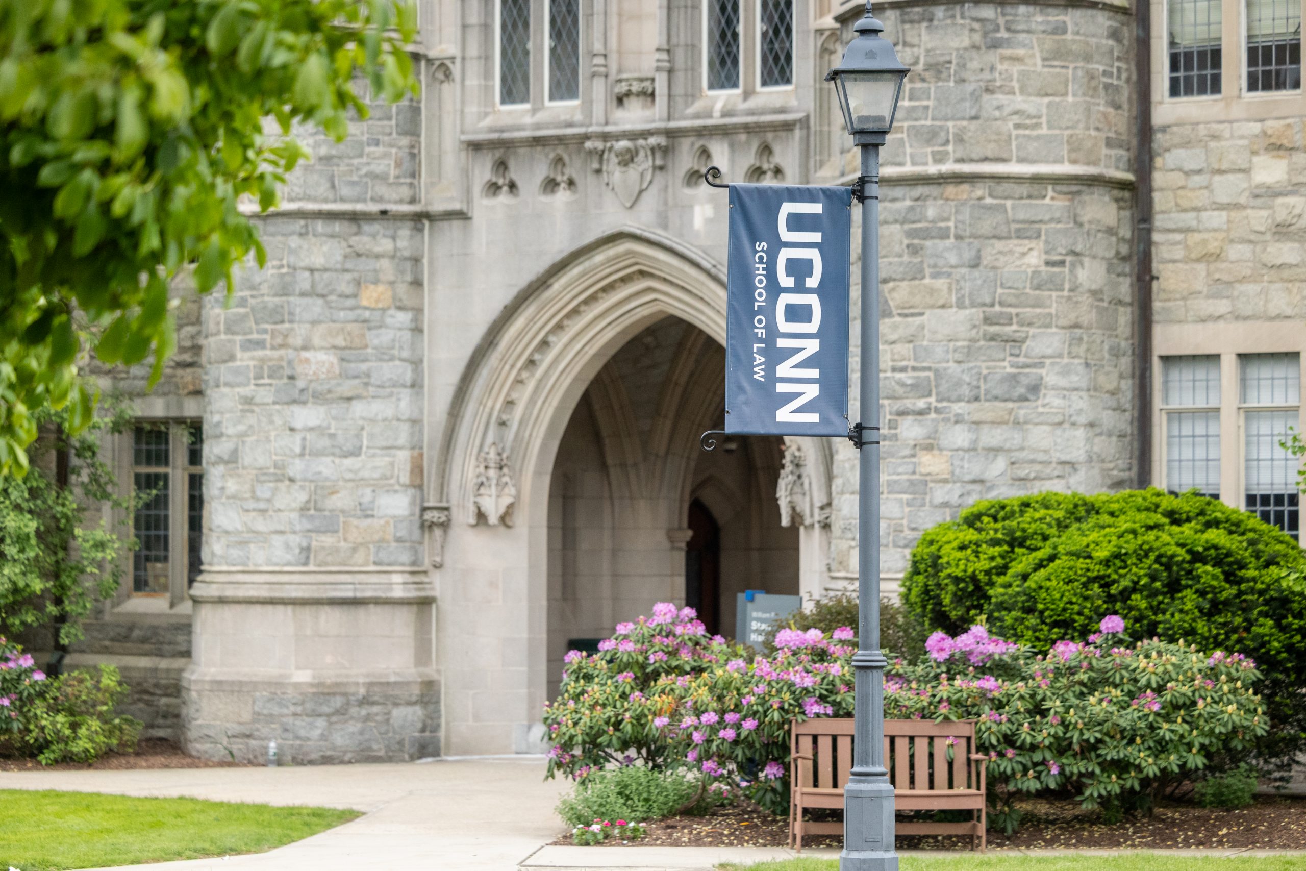 A UConn Law flag on a pole in front of a gray brick building with an arch, with green bushes and pink flowers.