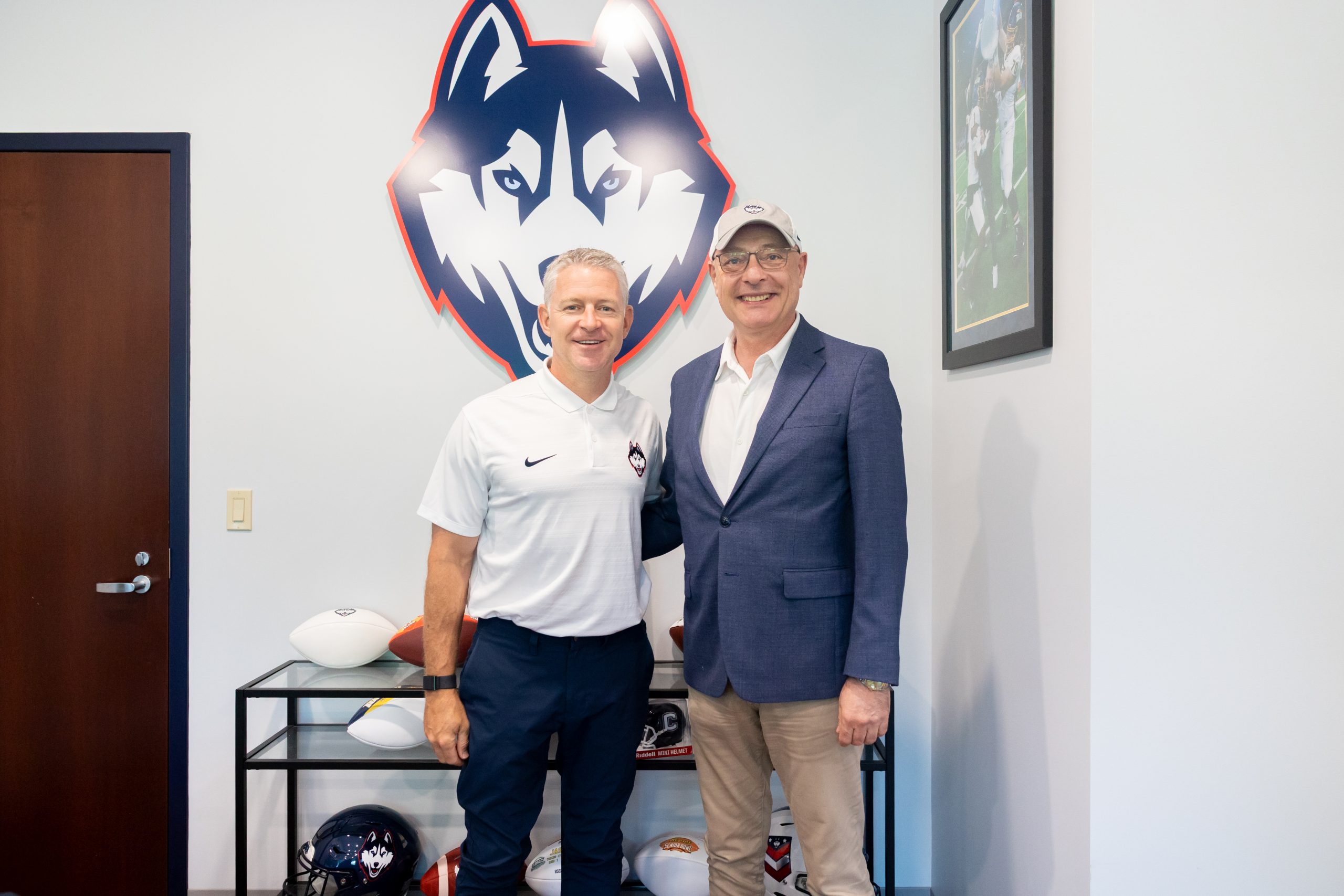 Two men posing for a photo in front of a Husky statue and a cart of footballs