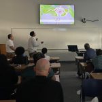 A student presents while gesturing at a powerpoint in front of a classroom full of people.