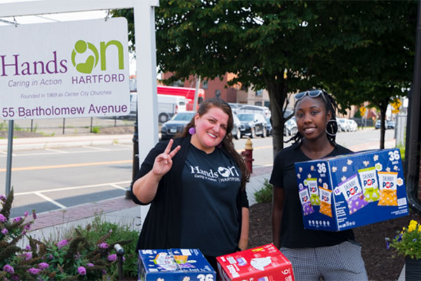 portrait two women holding donated goods next to Hands On Hartford sign
