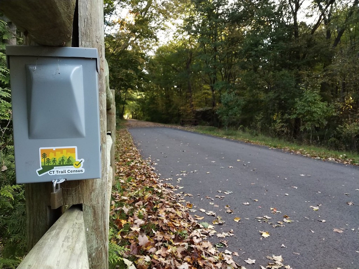 A paved trail surrounded by trees and undergrowth alongside a wooden fence. On the fence is a gray box with a Connecticut-shaped sticker on it.