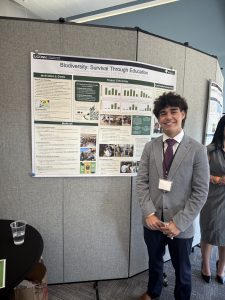 A student in a gray suit smiles by a research poster