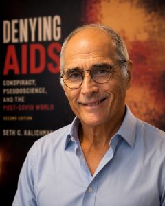 A man with glasses and a blue shirt smile at the camera with the words, "Denying AIDS" in large typeface behind him.