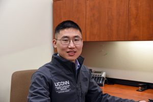 A man sits in a chair next to a desk wearing a UConn jacket.