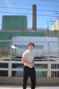 A student stands outdoors in front of an industrial power plant facility.