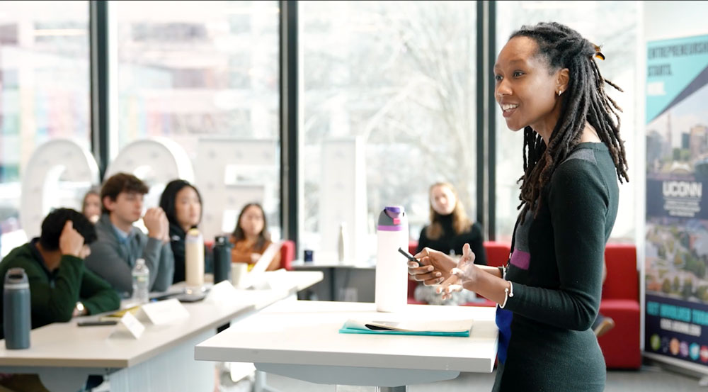 A woman is speaking to a group of people in a room full of windows.