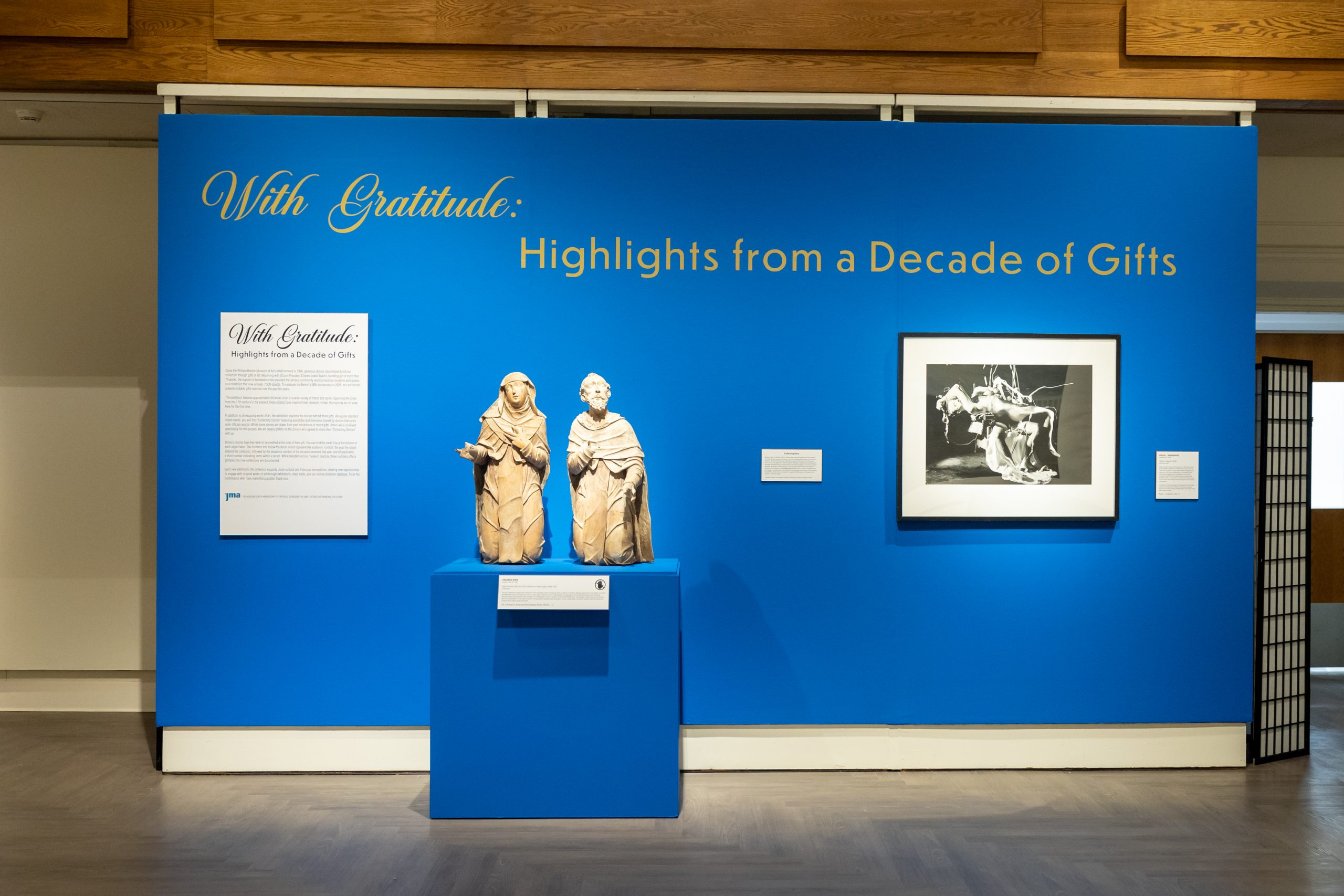 A wall painted blue with the words With Gratitude: Highlights from a Decade of Gifts on it along with a photo and sculptures
