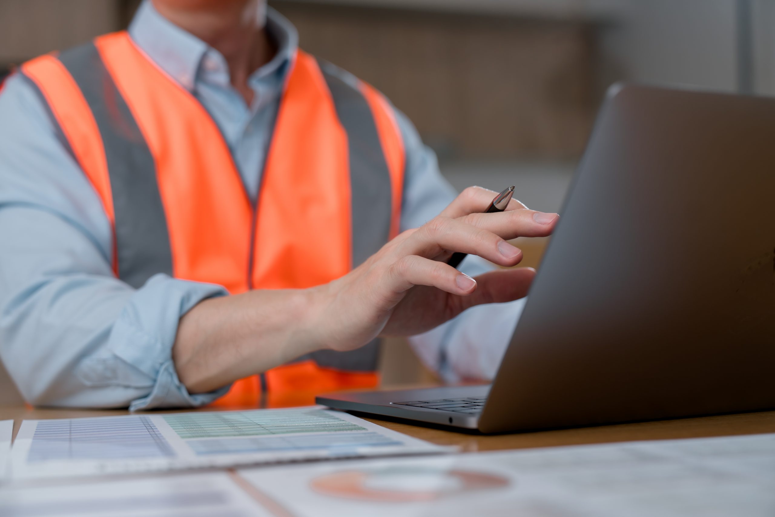 Close-up of an industrial worker wearing high visibility safety vest using laptop computer and reviewing documents in office or construction workplace environment.
