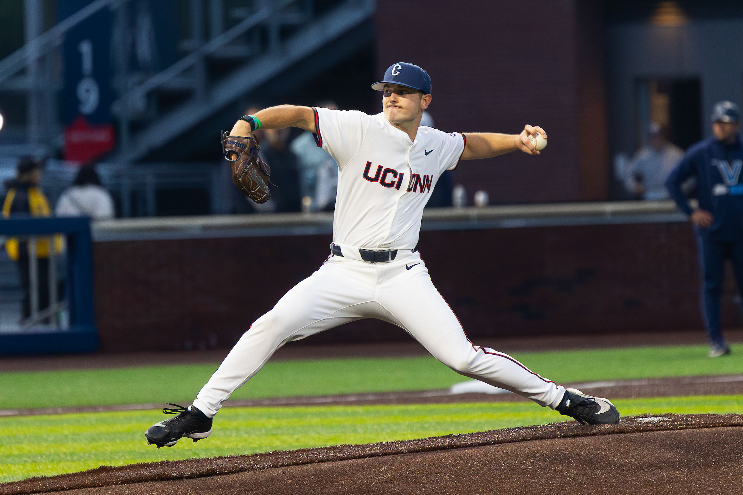 A baseball pitcher in the midst of throwing a pitch with "UConn" on a white jersey and a blue hat
