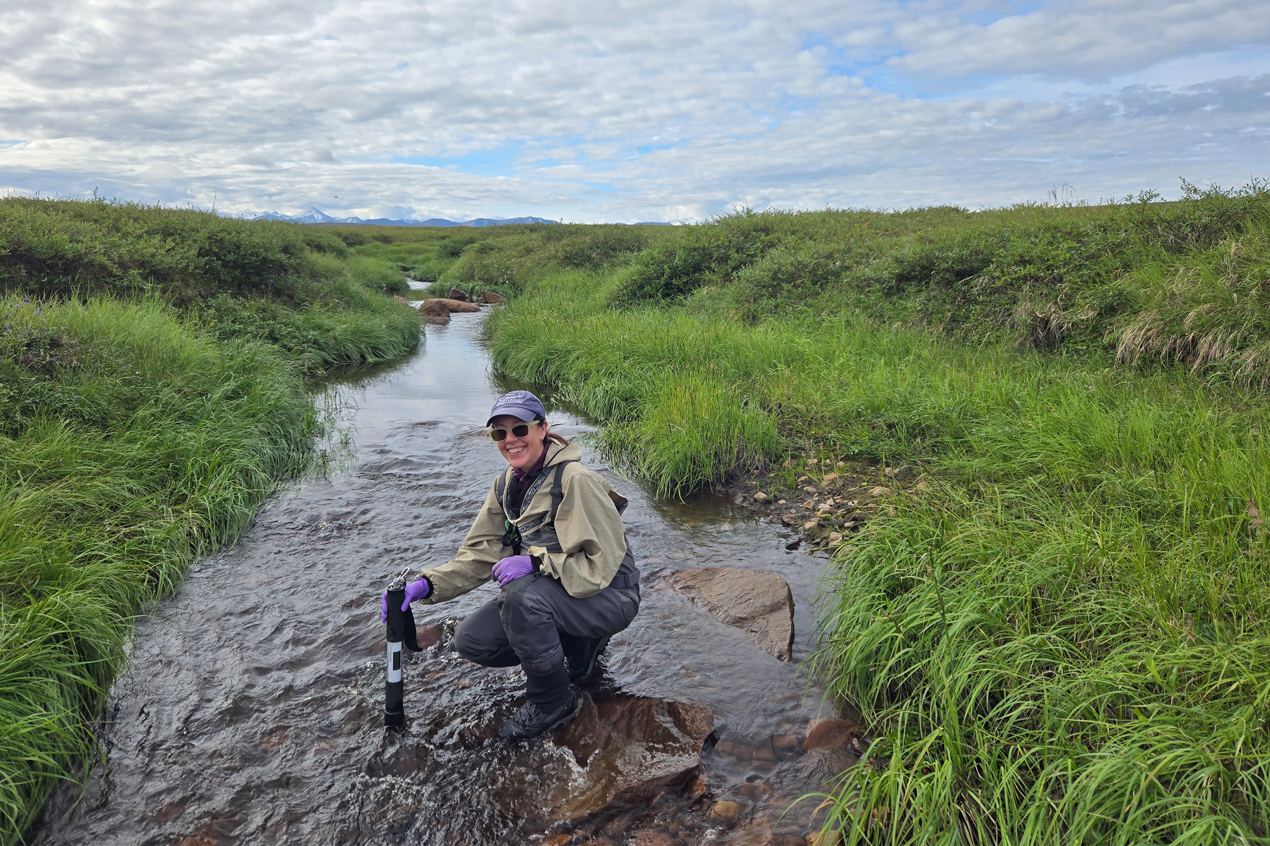 Drake_Benthotorch_UCTedit A person sitting next to a river holding an instrument to measure nutrients in the water.