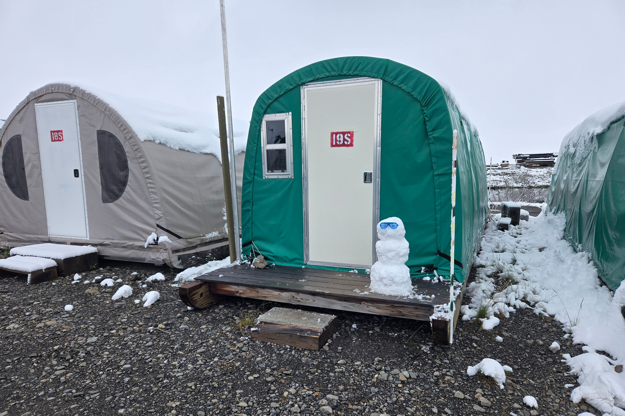 Drake_July_UCTedit Three tents covered in snow, one has a snowman in front of it with sunglasses on.