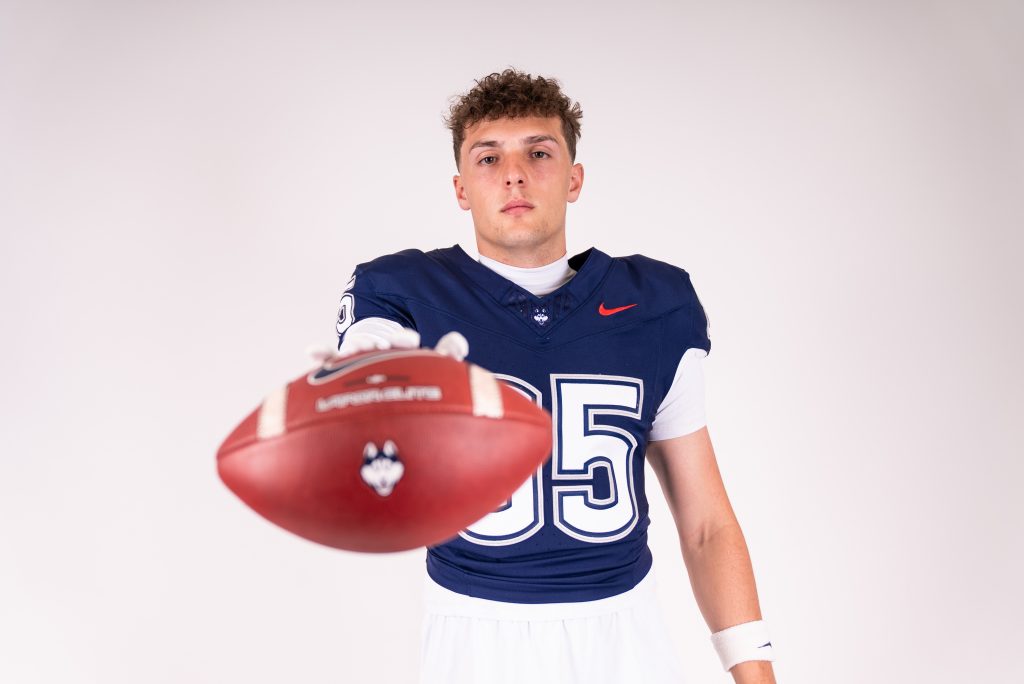 A young man in a blue football jersey holds a football toward the photographer.