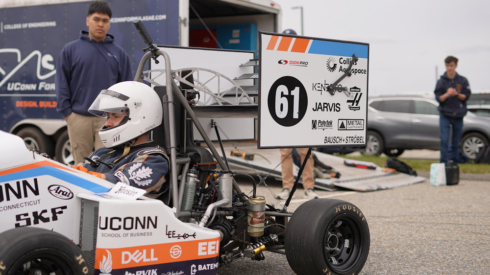 UConn Formula SAE race car sits in a paddock area with a helmeted driver in the cockpit