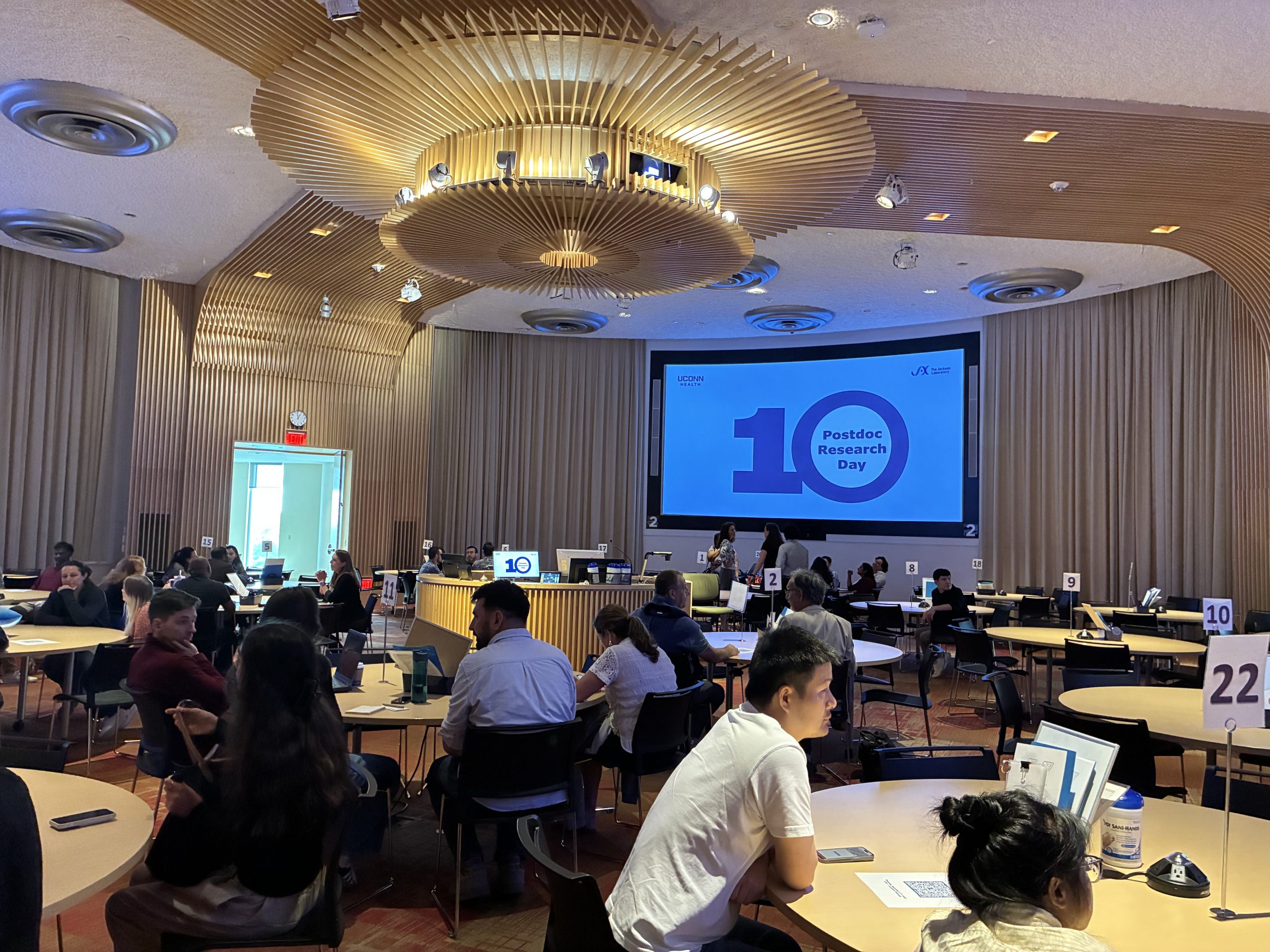 A room full of people at tables watches a large monitor with the number 10 on it.