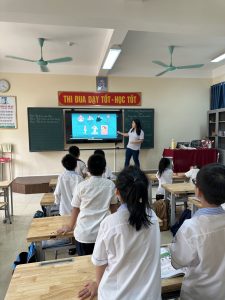 Lynna Vo stands at the front of a classroom and points at a screen while elementary school students look on.