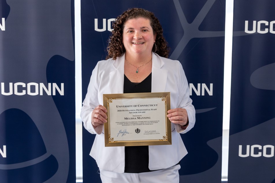 A woman in white holds an award certificate in front of a wall with UConn logos.