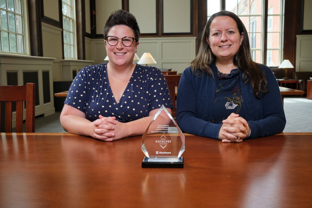 Two women sit side by side in front of a crystal award.