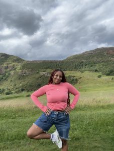 A woman with long red hair, a pink long-sleeved top, and denim shorts poses on one leg in front of a green rolling hill.