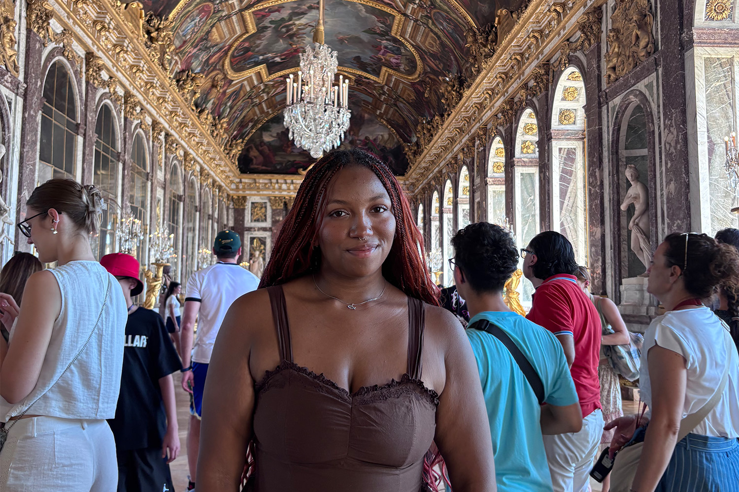 A woman with long hair and dressed in a brown sundress poses in an ornate French palace with a crowd of tourists around her.