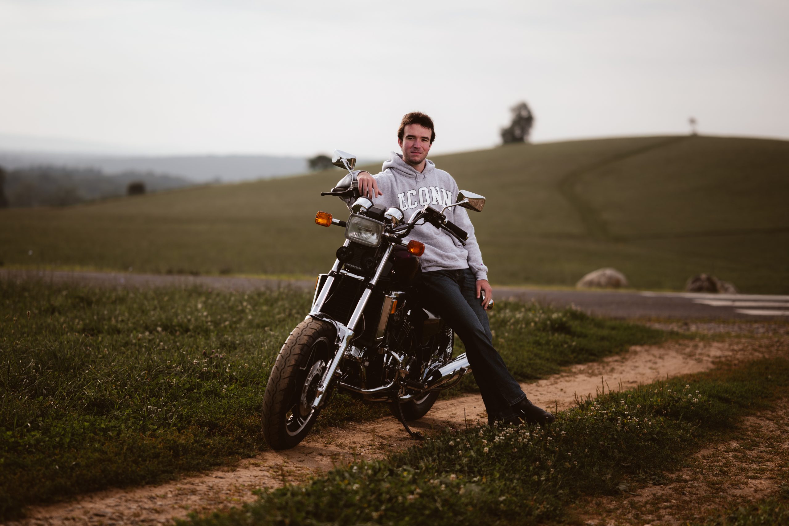A young man in a gray sweatshirt leans on a motorcycle on Horsebarn Hill on an overcast day.